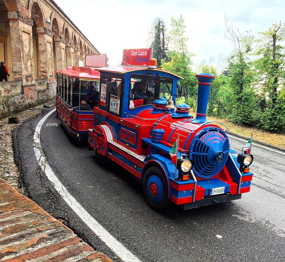 Red and blue tourist train driving on a curved road near an arched brick wall and greenery in the background.