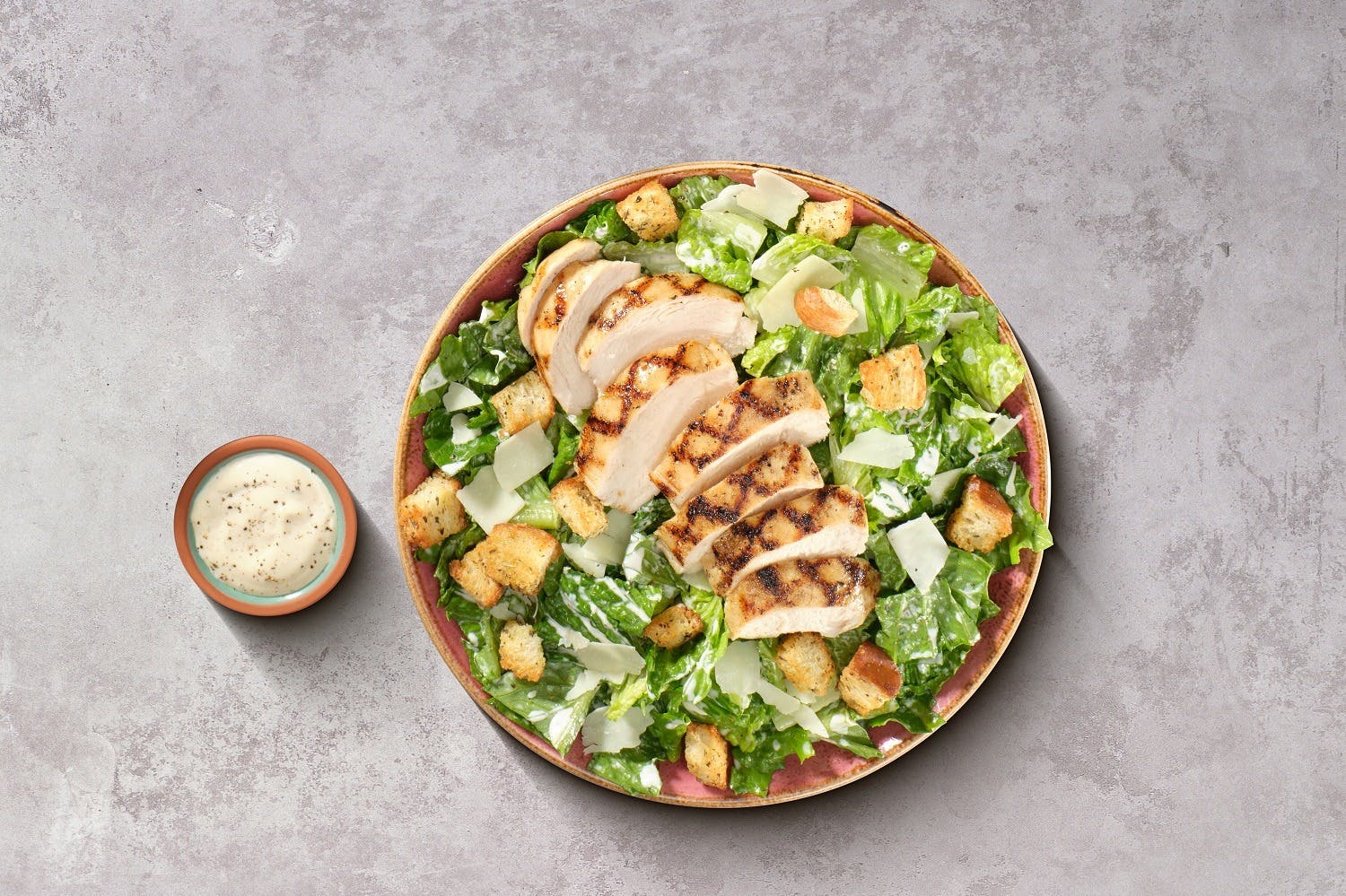 A Caesar salad with grilled chicken slices, croutons, and Parmesan, alongside a side of creamy dressing in a small bowl.