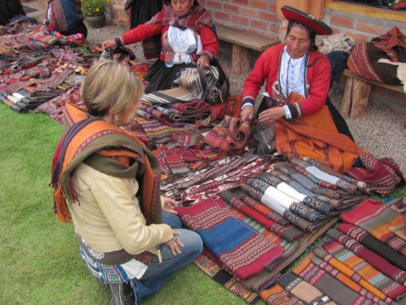 Two women in traditional dress sell colorful textiles on the ground while a woman kneels and examines the items.