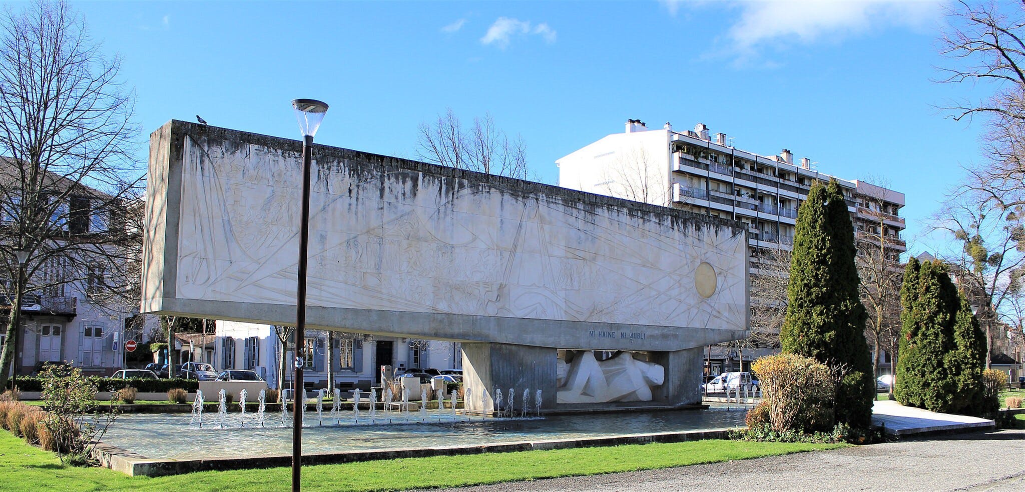 反射するプール、周囲の木々、茂み、近くの住宅の上に刻まれたデザインのコンクリートモニュメント。