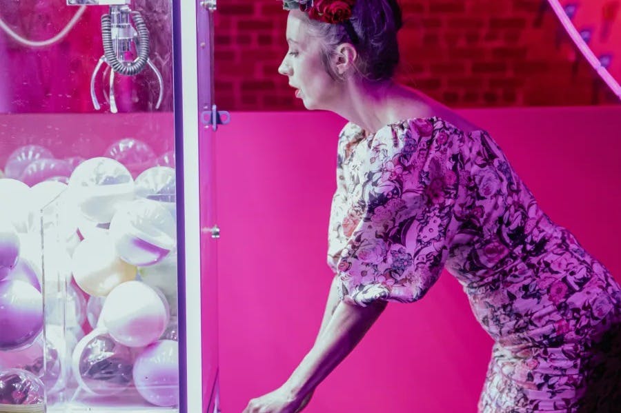 꽃무늬 드레스와 화관을 쓴 여성이 분홍색 배경에 흰색 공으로 채워진 발톱 기계를 작동합니다.