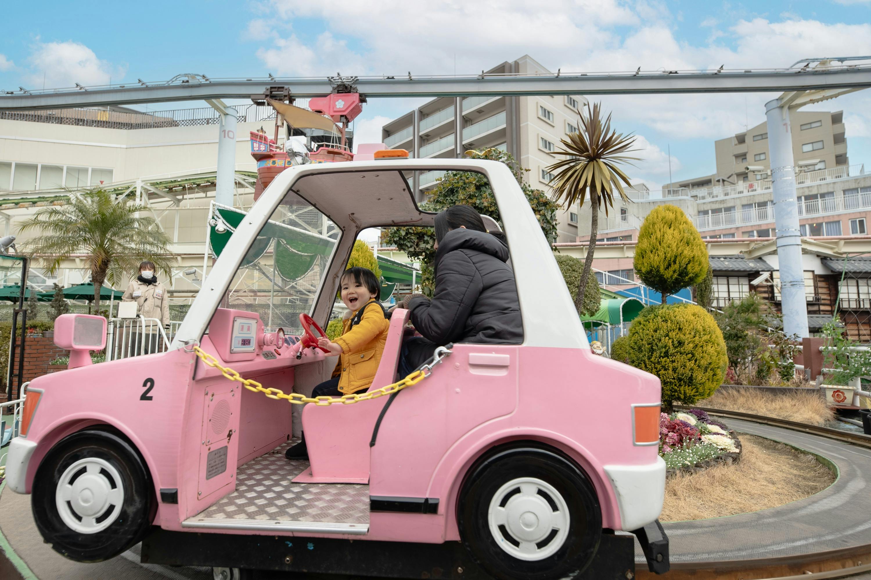 A child in a yellow jacket and an adult ride in a pink toy car at an amusement park, with a cityscape in the background.