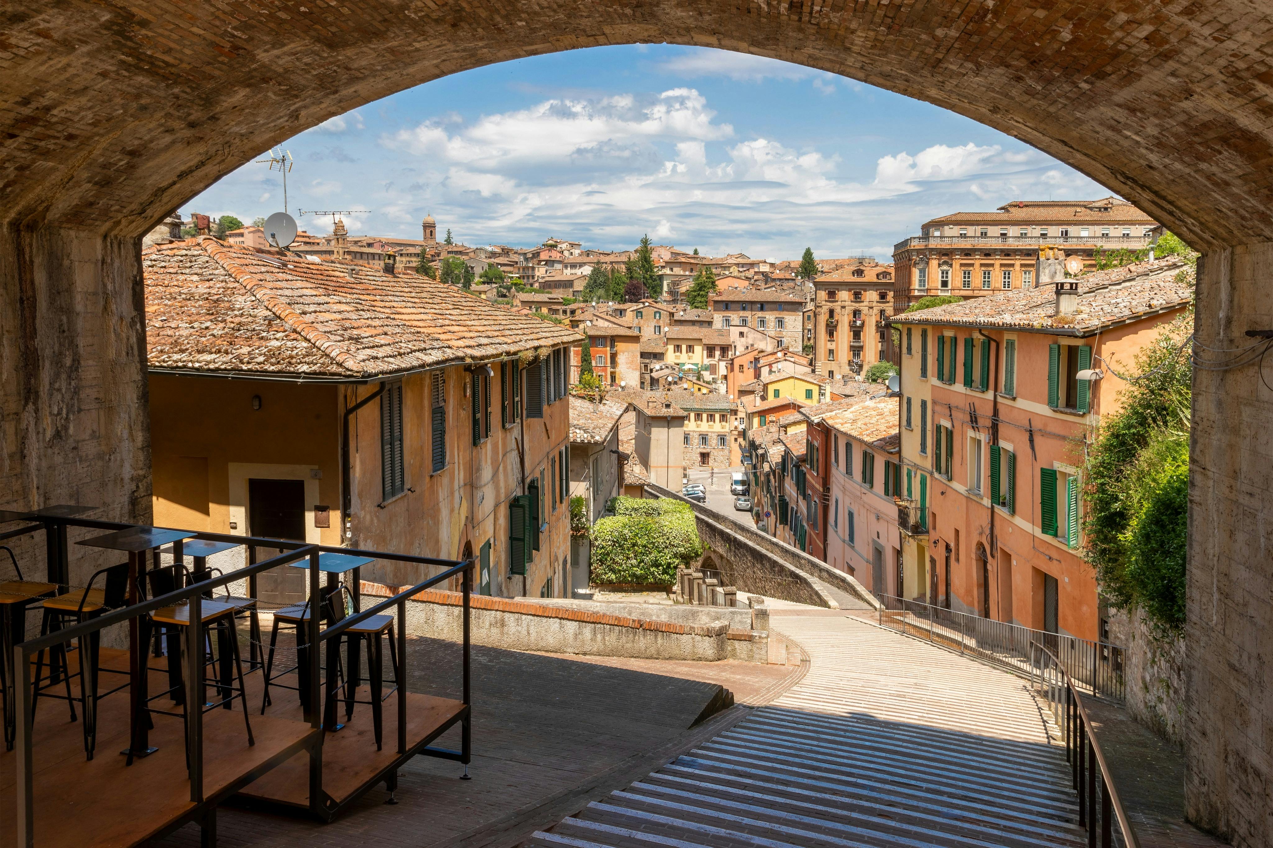 Una veduta panoramica di una città storica italiana con edifici dai tetti in tegole, vista sotto un arco in pietra, con una terrazza in primo piano.