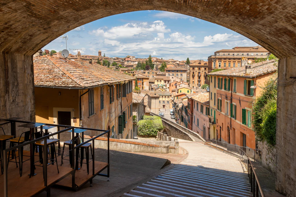A scenic view of a historic Italian town with tiled-roof buildings, seen under a stone archway, with a terrace in the foreground.