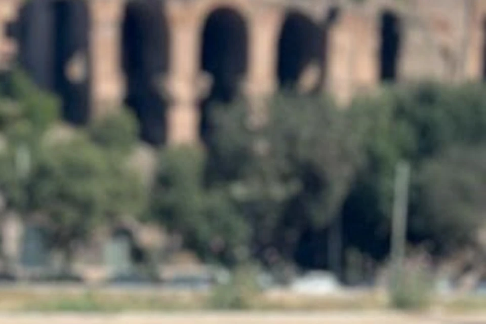 Woman in a helmet and sunglasses, sitting on a bicycle, with ancient ruins and a clear blue sky in the background.