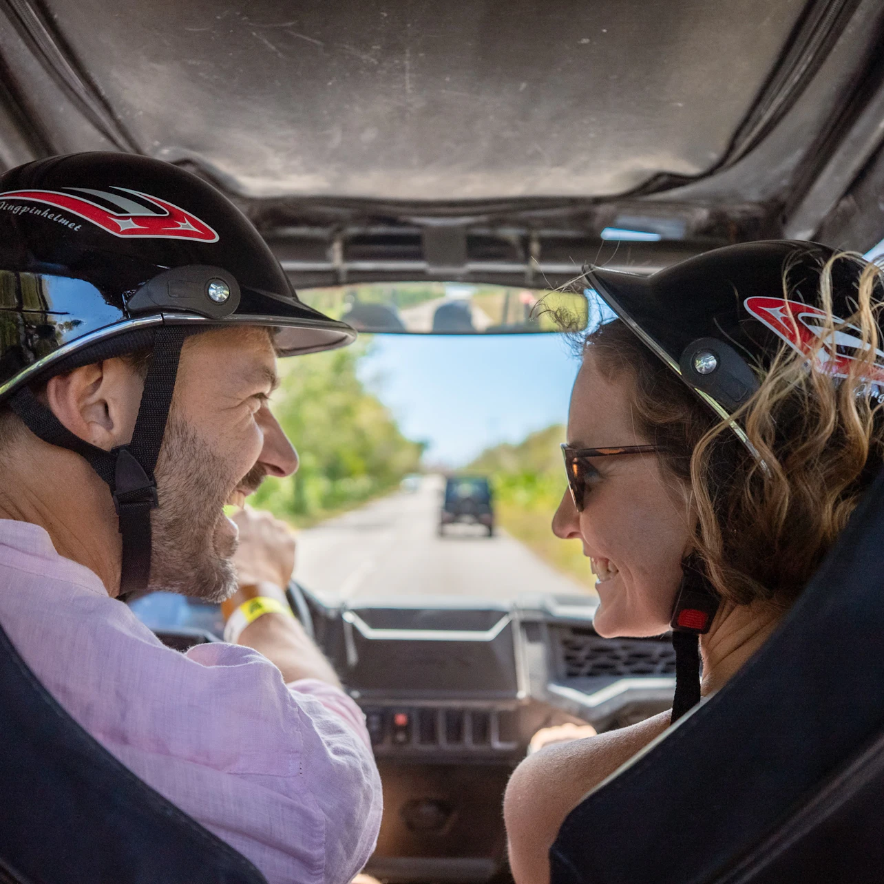 Two people wearing helmets and sunglasses ride in an open vehicle on a sunny road through a lush, green landscape.