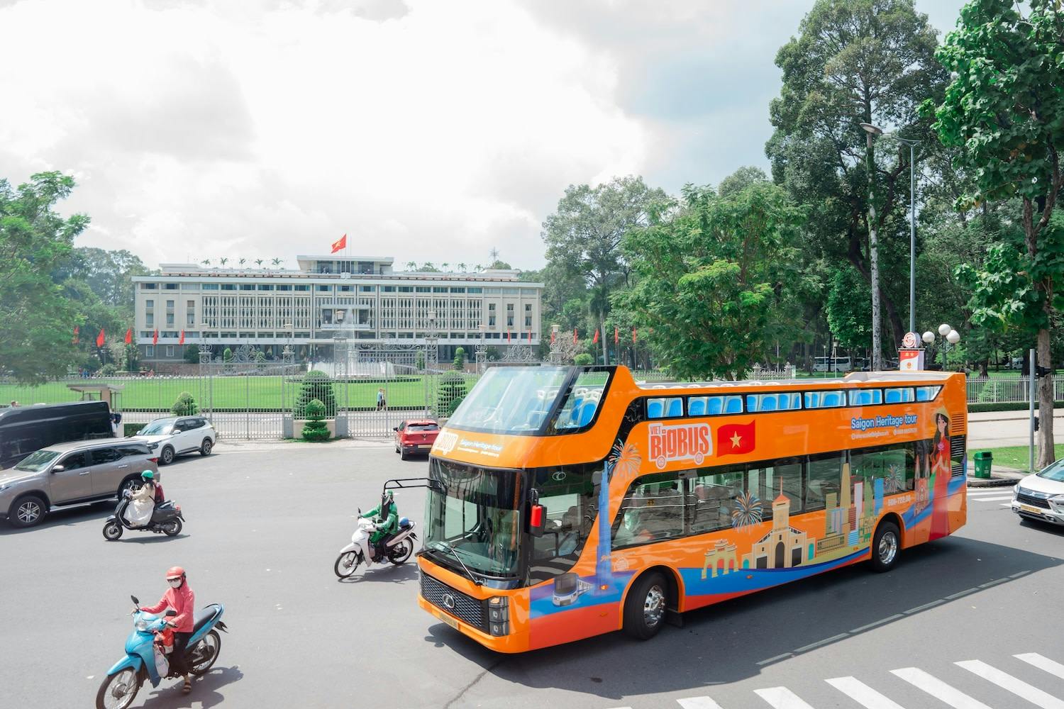 An orange double-decker tour bus on a street near a large building with a flag and green lawn, surrounded by trees.