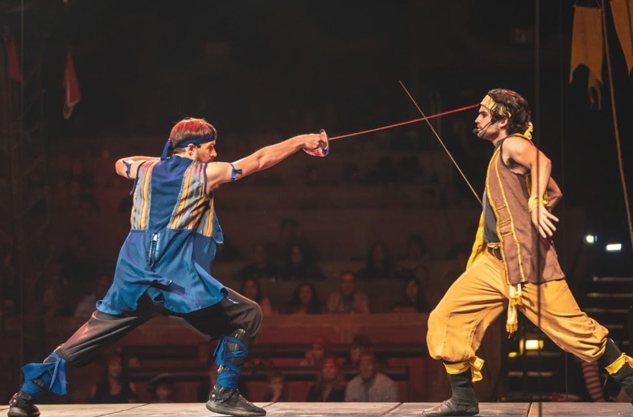 Two performers engage in a dramatic sword fight on stage, with an audience watching in the background.