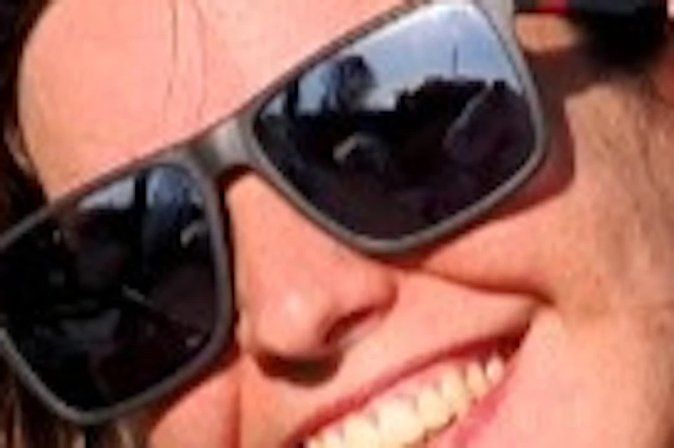 Two smiling people wearing sunglasses pose with arms raised inside a green and black off-road vehicle under a clear blue sky.