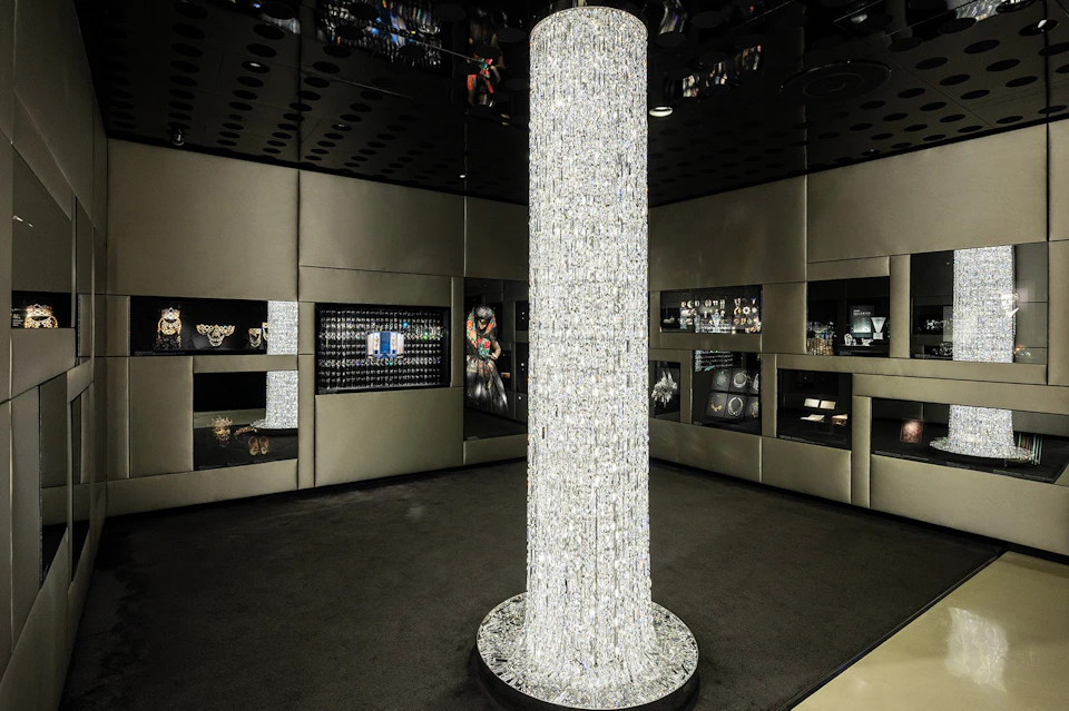 A luxurious showroom featuring a tall, cylindrical crystal chandelier surrounded by display cases with jewelry and accessories.