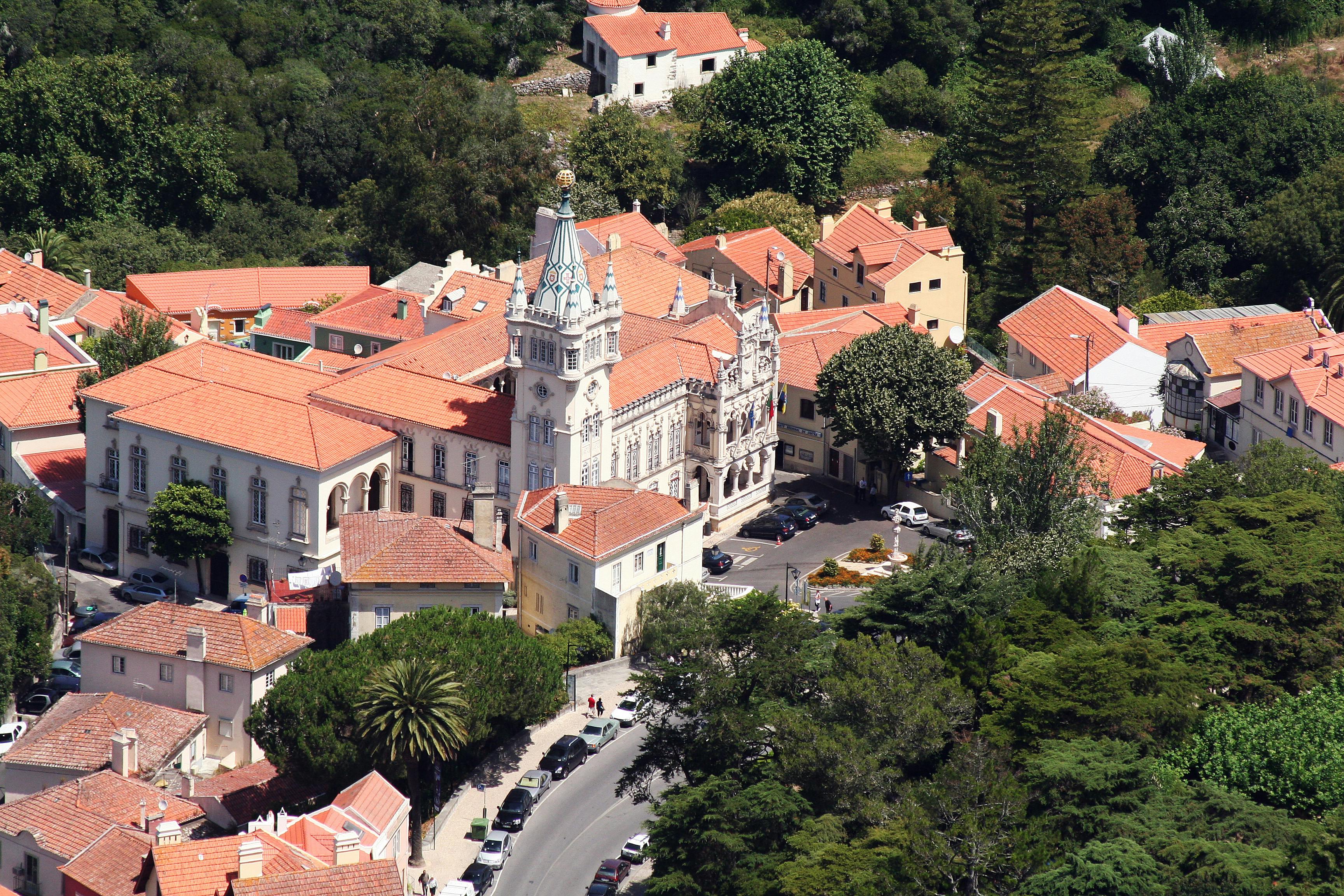 Εναέρια άποψη ενός ιστορικού κτιρίου με πύργο, που περιβάλλεται από σπίτια με κόκκινες στέγες και δέντρα σε μια ηλιόλουστη μέρα.