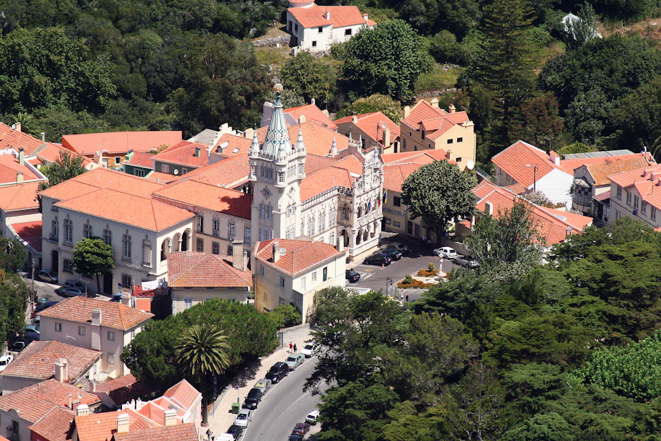 Aerial view of a historic building with a tower, surrounded by red-roofed houses and trees on a sunny day.