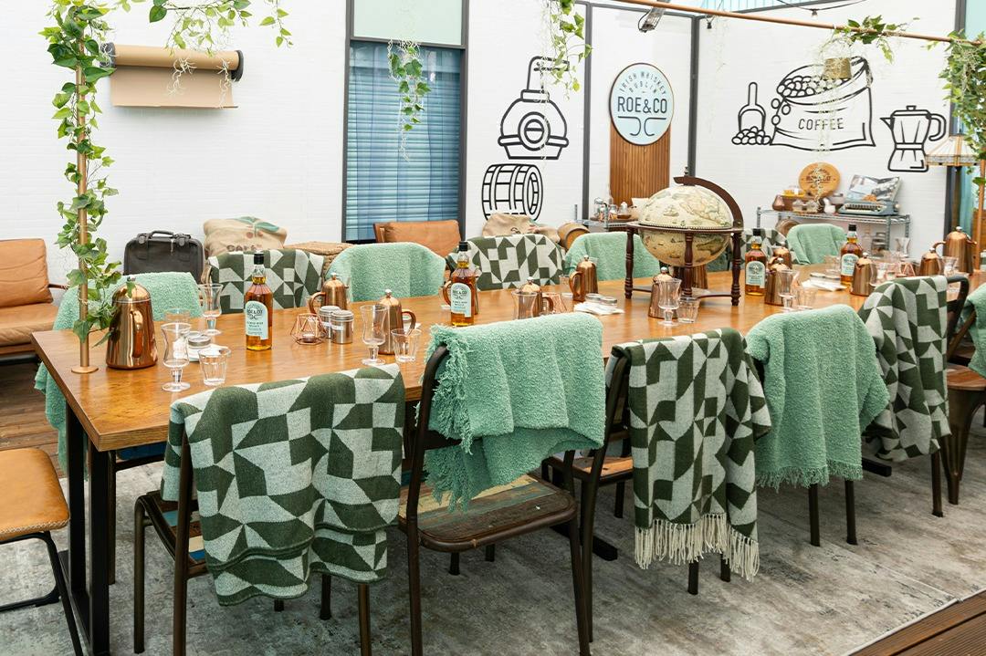 A decorated table set with bottles, glasses, and green patterned blankets draped over chairs, in a room with wall illustrations.