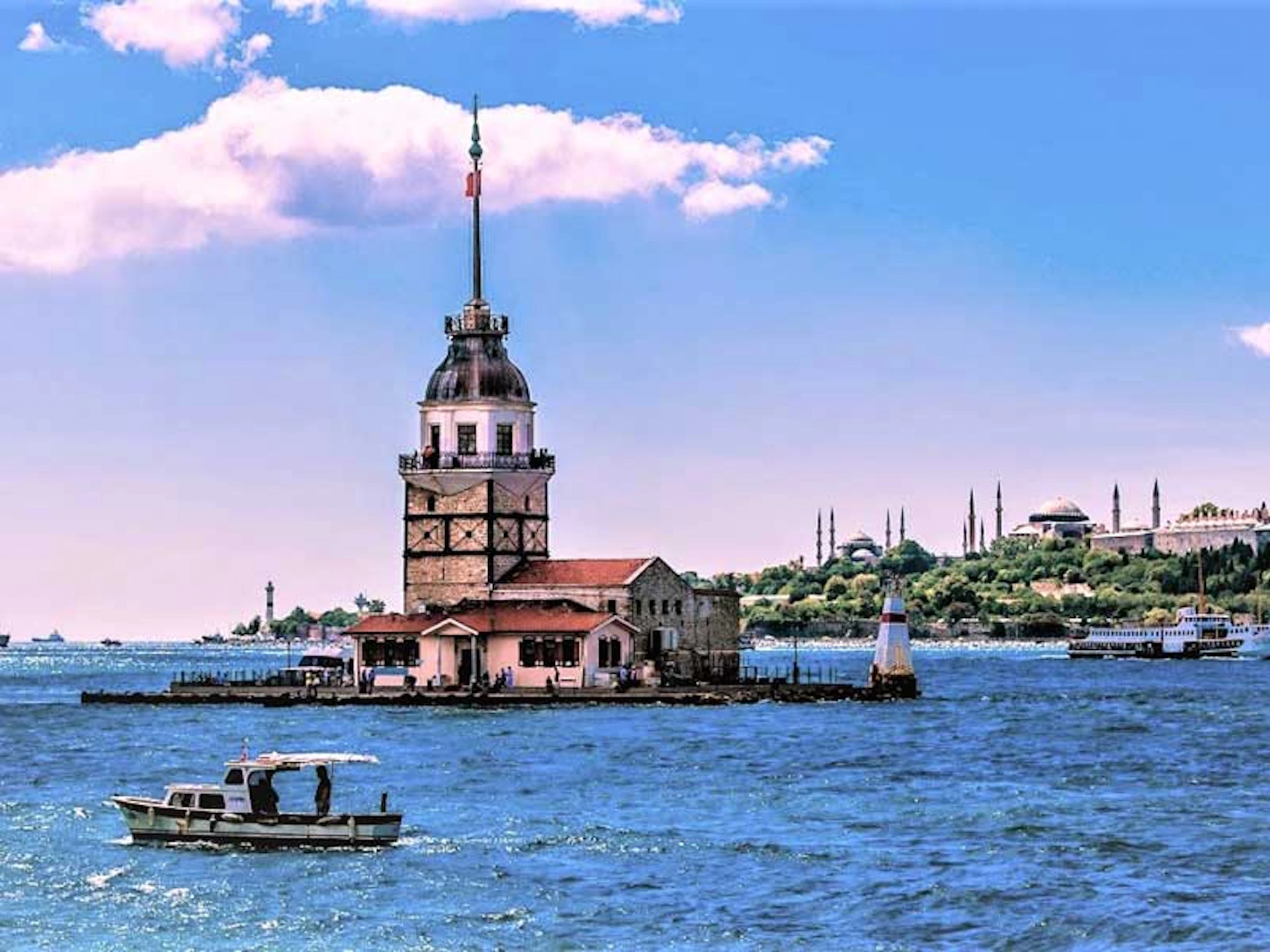 A historic tower on a small island in a body of water with a boat in the foreground and a cityscape in the background under a blue sky.