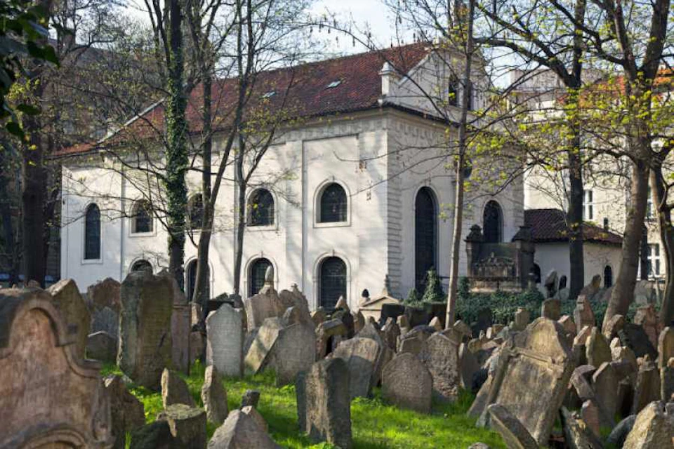 Old gravestones scattered in a grassy cemetery, with a historic white building featuring arched windows and a red-tiled roof in the background.