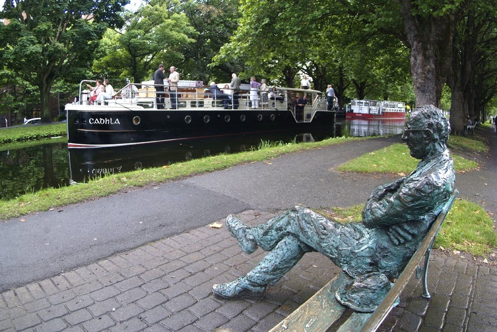 一尊雕像坐落在运河附近的长凳上，一艘载有乘客的船停泊在那里。 后面是树木和一条铺好的小路。