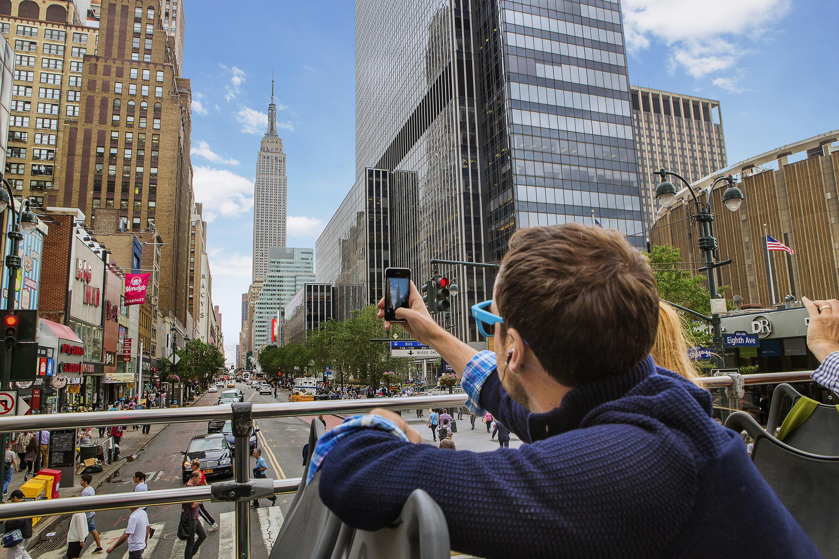 En person tar ett foto av Empire State Building från en öppen buss på en livlig gata i New York City.