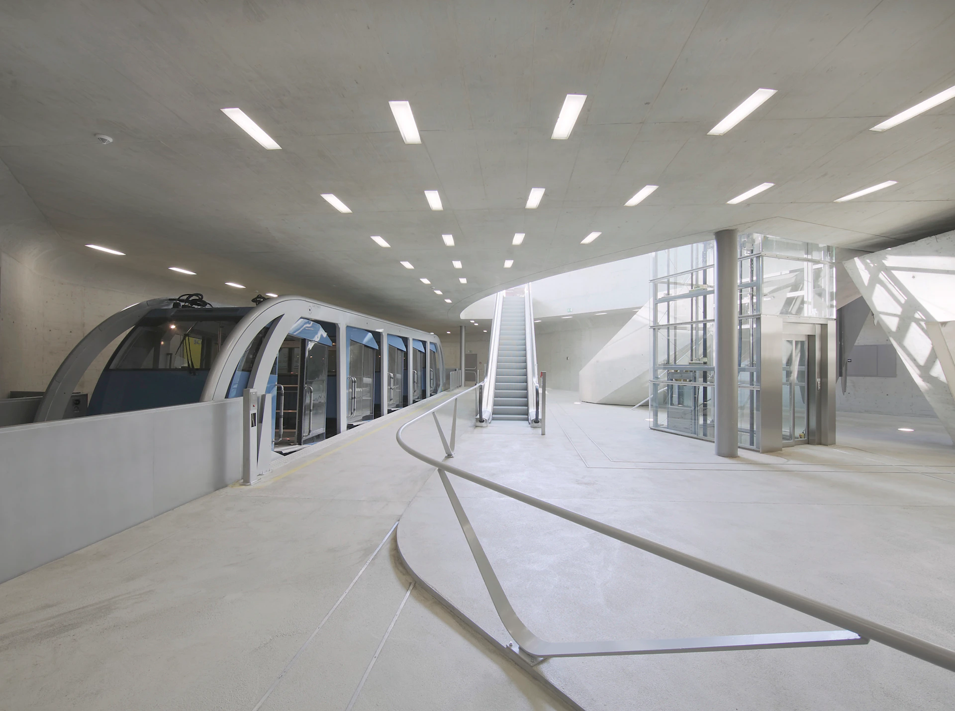 Modern indoor train station with a sleek monorail, escalators, and glass-enclosed elevator. Bright lighting from ceiling fixtures.