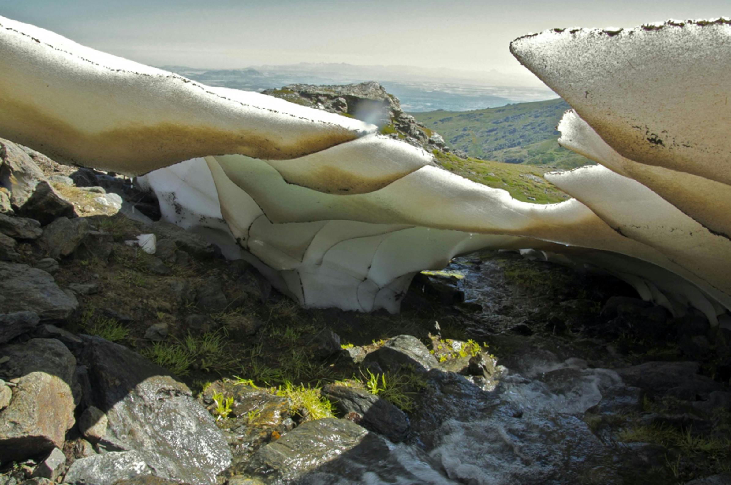 Large sheets of ice over a rocky stream with patches of grass, set against a hilly landscape under a clear sky.