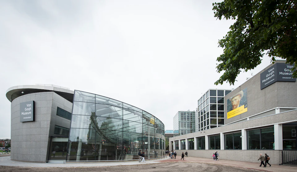 Modern glass building with a curved facade, adjacent to a square building with grid windows. Few people are walking nearby.