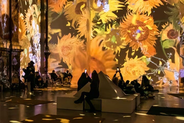 People seated in a room with large sunflowers projected on the walls, some taking photos with their phones.