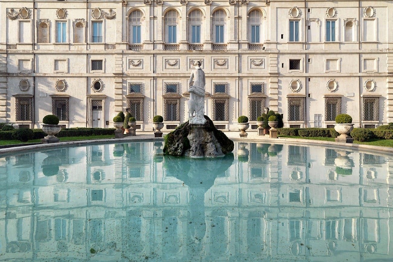 Statue in the center of a circular fountain in front of an ornate, classic-style building with manicured bushes on both sides.
