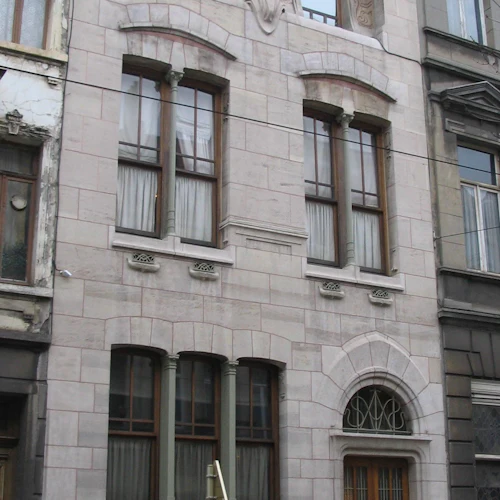 Trevånings stenbyggnad med välvda fönster och invecklade arkitektoniska detaljer. Trädörr på gatunivå.