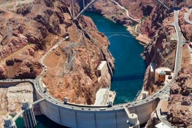 Aerial view of a large dam holding back a river, surrounded by rugged desert terrain and a bridge in the background.