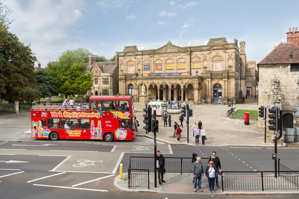 People walk near an ornate historic building. A red sightseeing bus and several pedestrians are visible. Traffic signals and crosswalks are in the foreground.