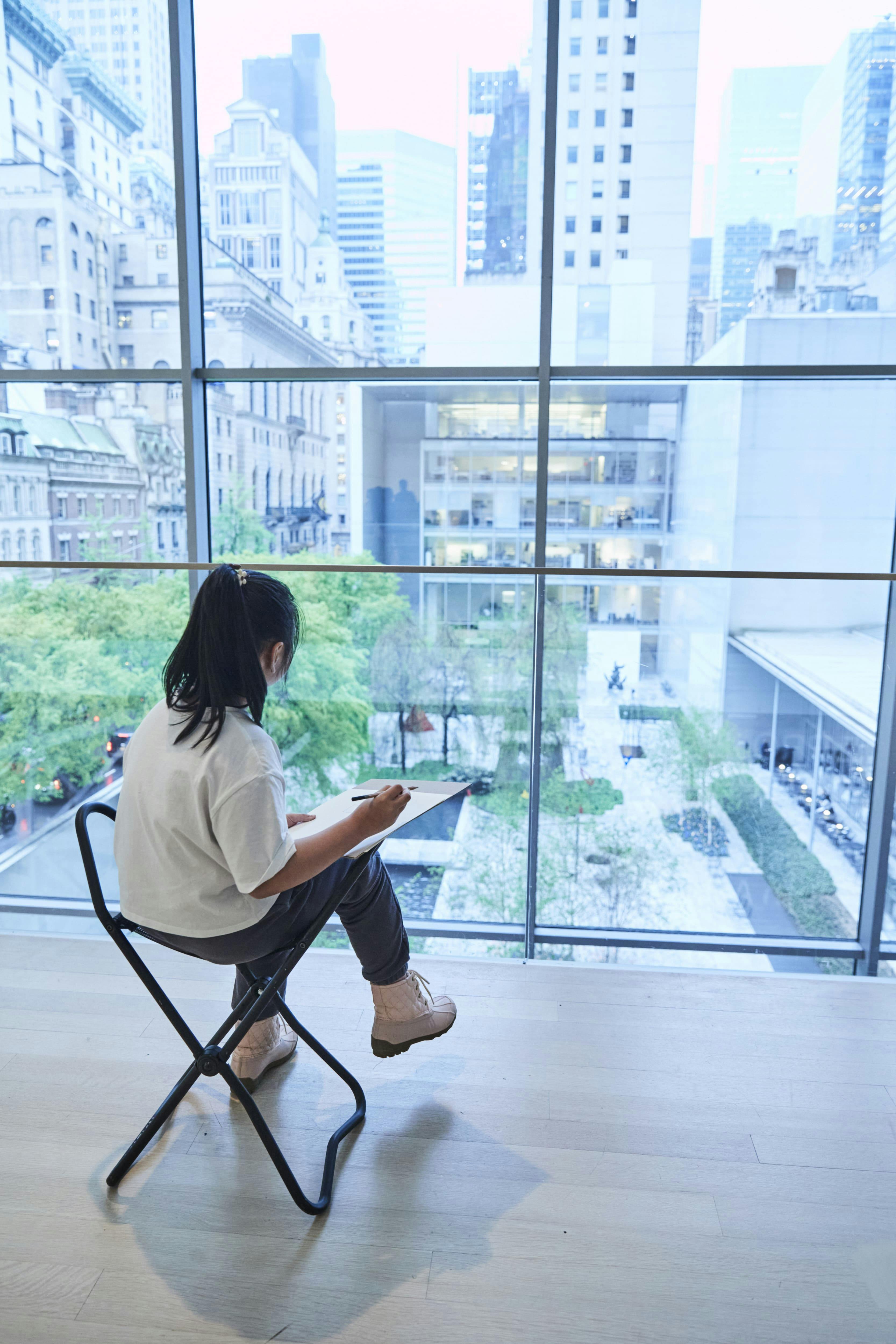 Člověk sedí na stoličce, kreslí na velkou podložku a dívá se na okno o velikosti stěny s výhledem na městské nádvoří se stromy a budovami.