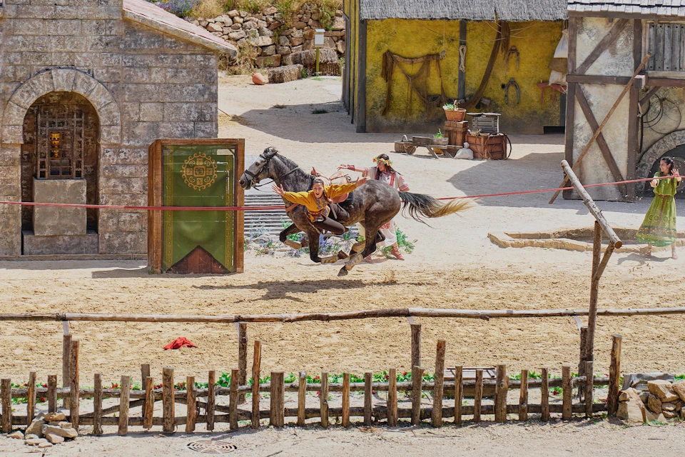 Two performers in medieval attire act out a scene with a horse in a sandy courtyard, surrounded by rustic buildings.