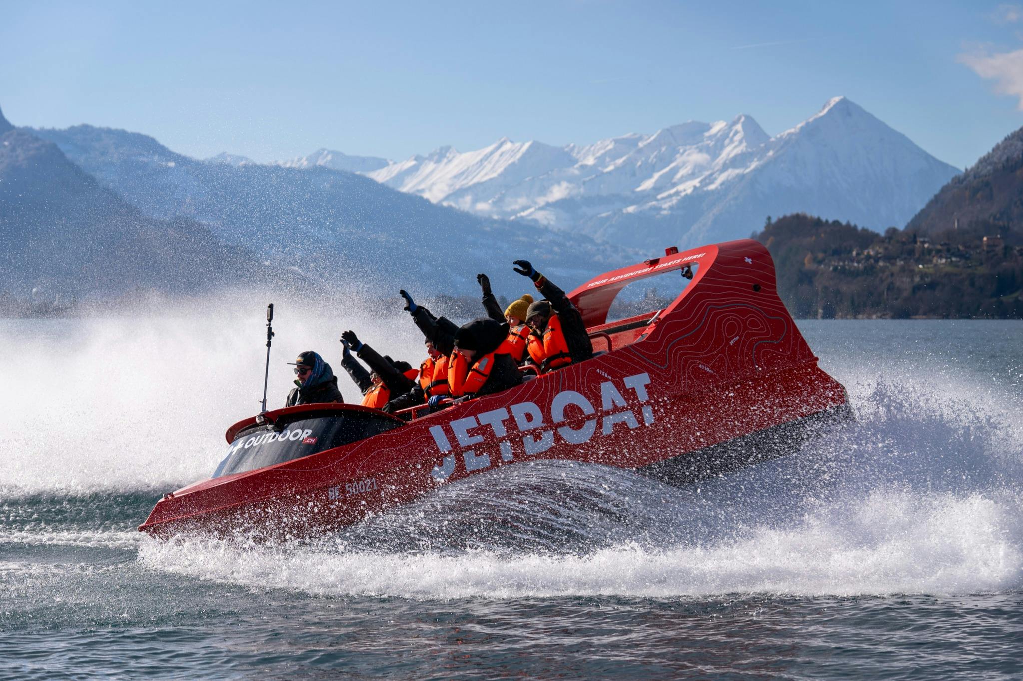A red jet boat labeled "JETBOAT" speeds across water, carrying passengers in life vests, with snow-covered mountains in the background.