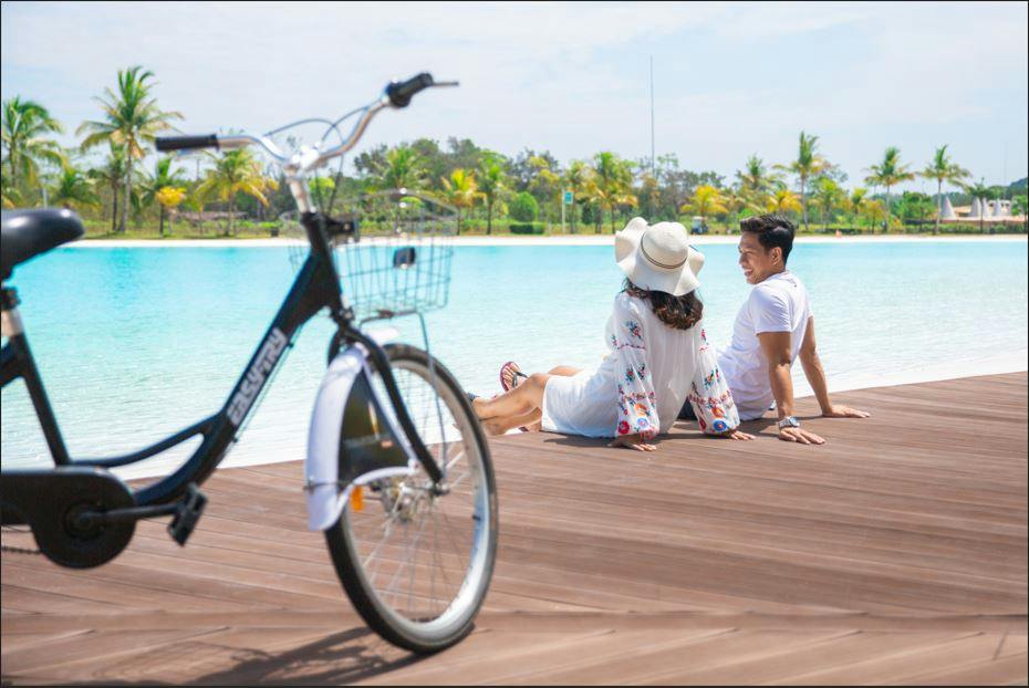 A couple sitting on a wooden dock by a tropical lagoon, with a bicycle in the foreground and palm trees in the background.