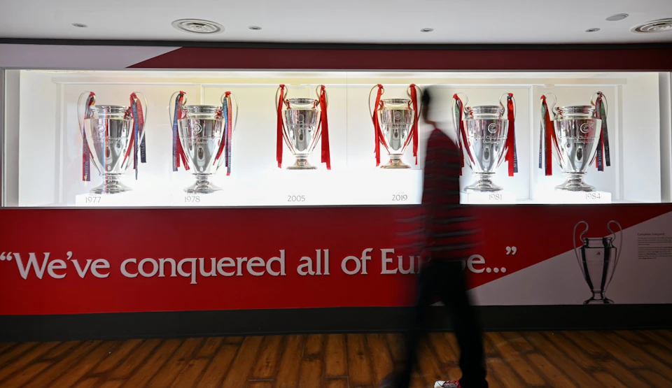 A person walks past a display of five silver trophies with red ribbons, each labeled with a year, against a red and white wall.