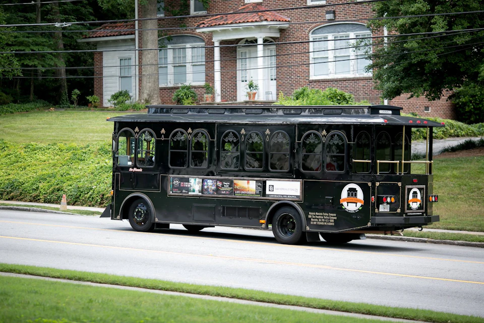 A black trolley bus is driving down a street in front of a red-brick house with white trim and arched windows.