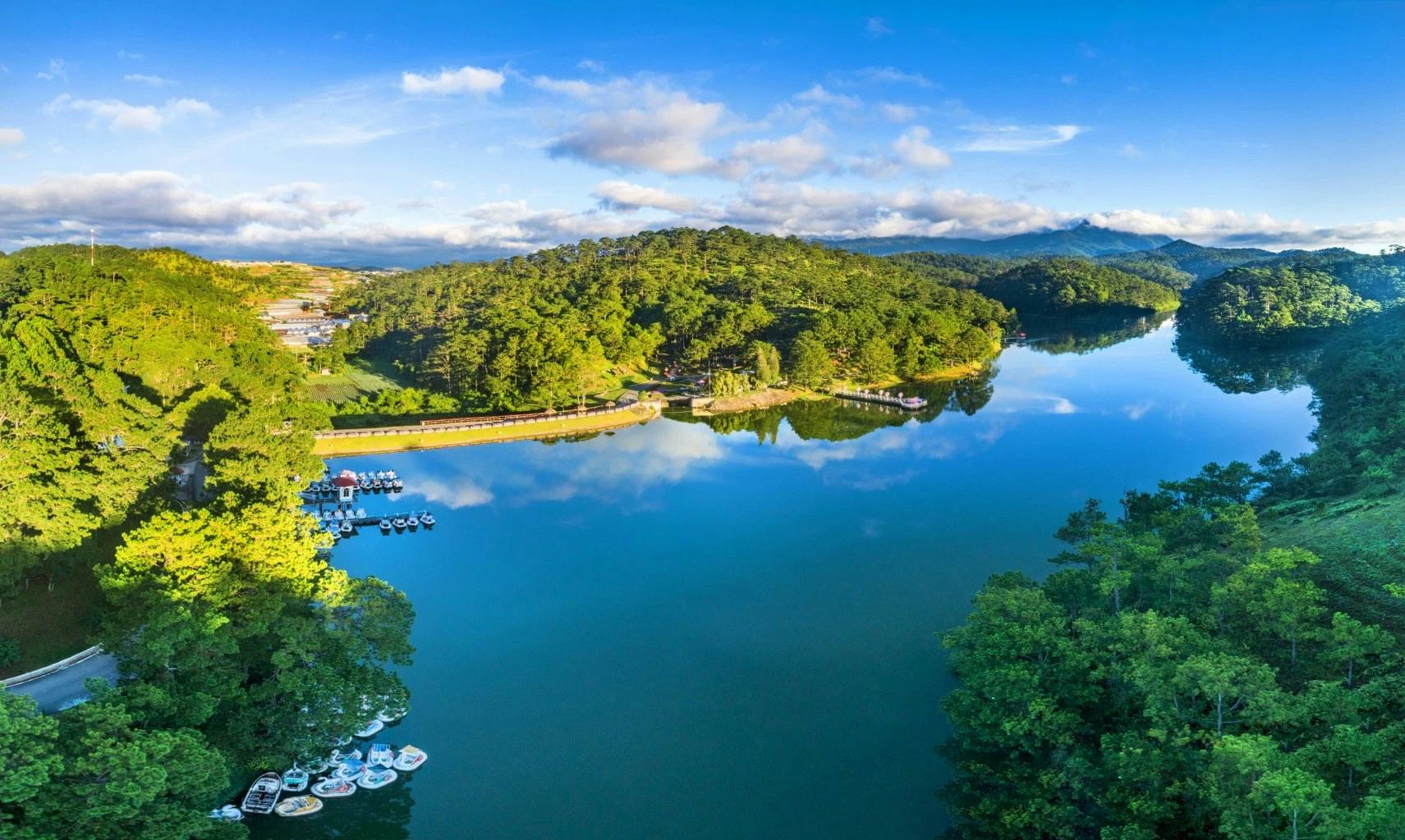 A serene lake surrounded by lush green forest under a blue sky with clouds, boats visible along the shore.
