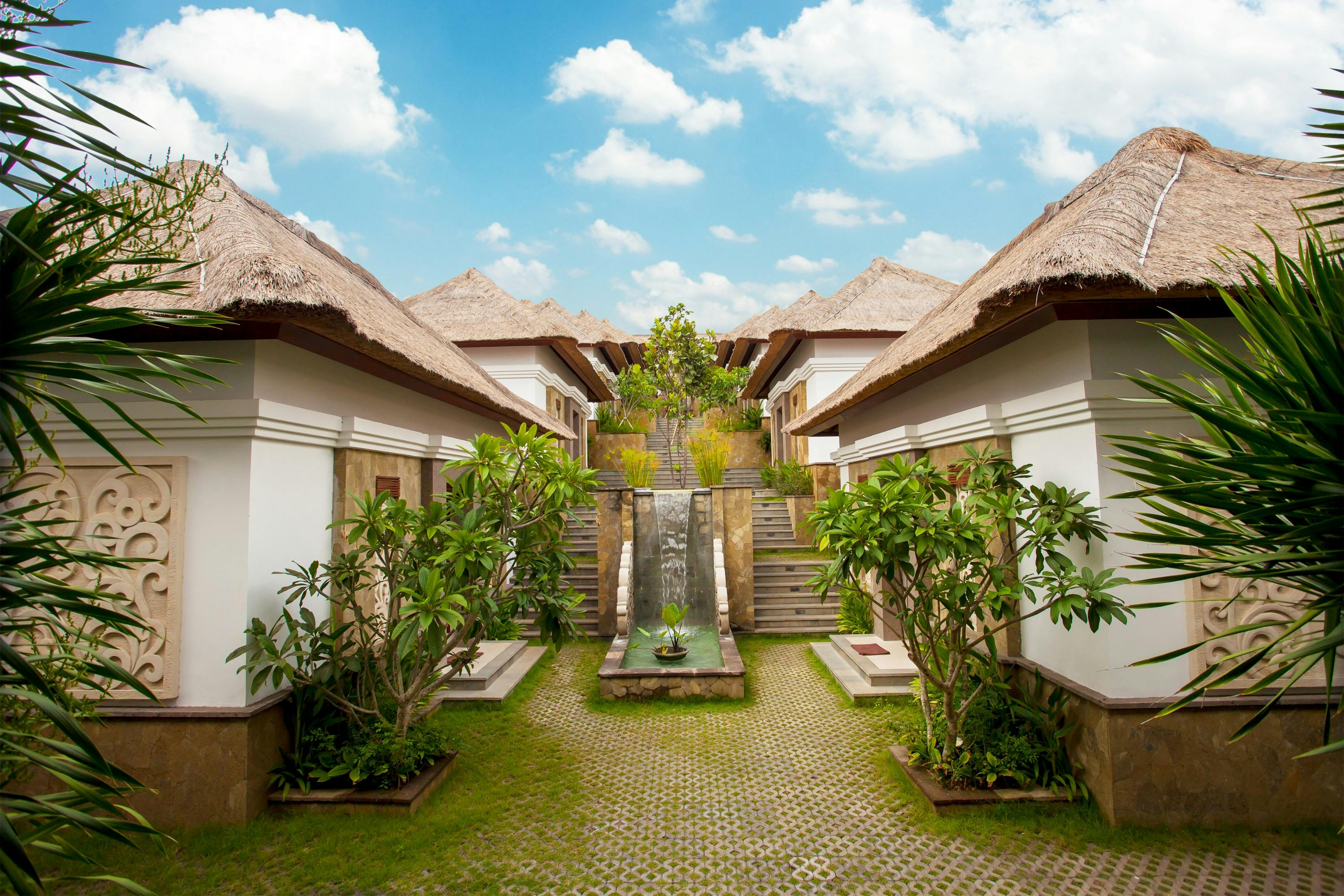 A courtyard with a water feature and lush plants, flanked by white buildings with thatched roofs under a blue sky.