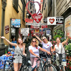 Fyra kvinnor med cyklar ler och vinkar i en smal, färgstark gränd prydd med många italienska skyltar och dekorationer.