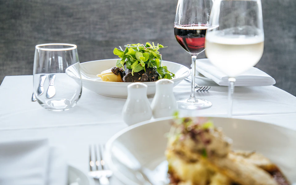 Plated dishes on a table with glasses of red and white wine, salt and pepper shakers, and a white tablecloth.
