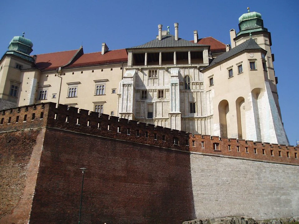 A large, historic building with a red roof and white walls behind a tall, brick retaining wall under a clear blue sky.