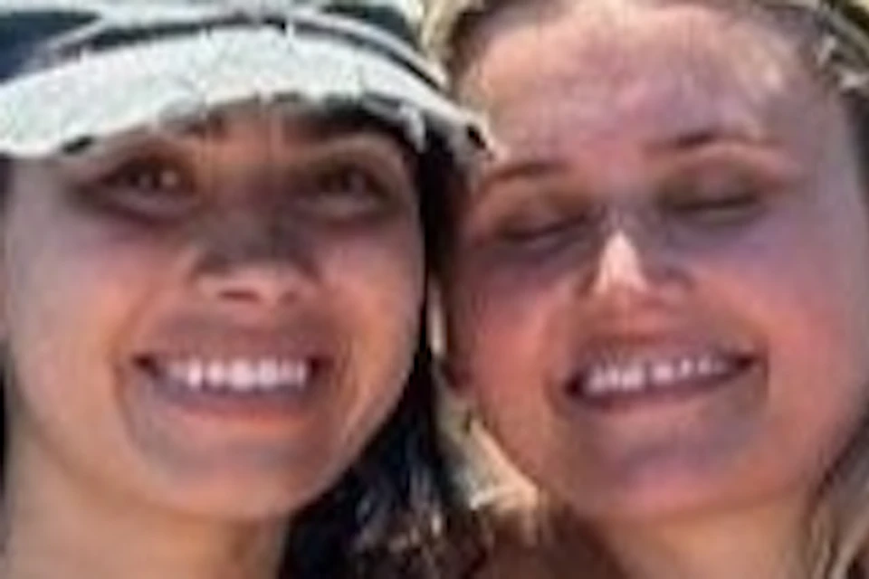 Two women in swimsuits pose together on a boat with a rocky coastal arch formation in the background under a clear sky.
