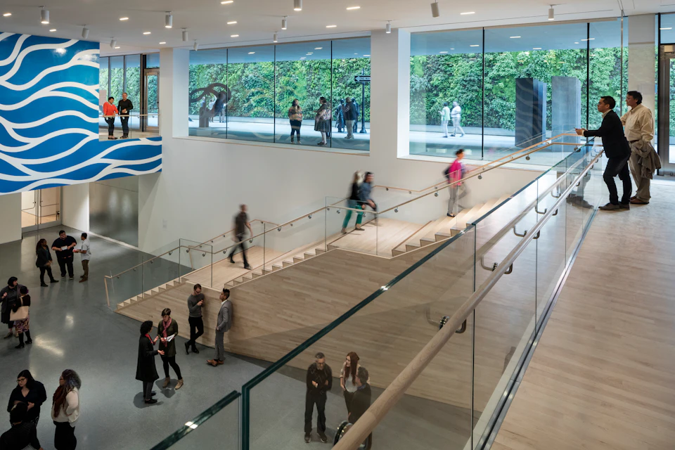 Modern museum interior with glass walls, indoor garden view, visitors on steps and walkways, and a few sculptures in the background.
