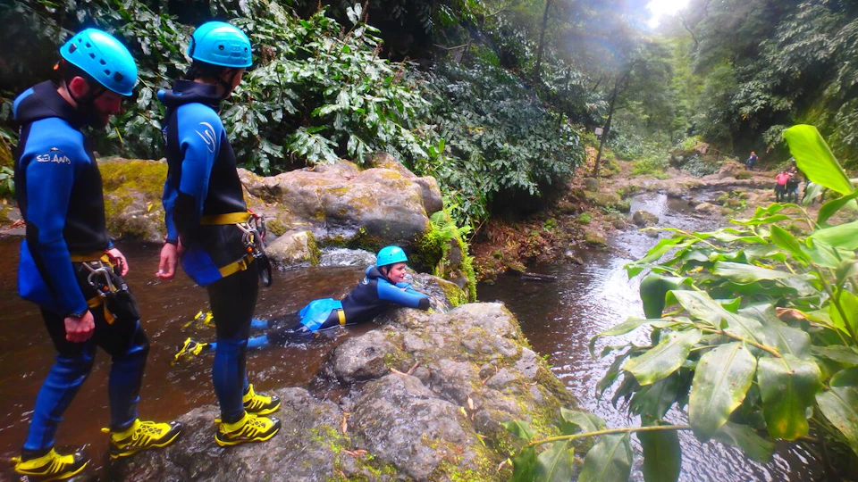 Two people wearing wetsuits and helmets, one resting on rock beside a forest stream, surrounded by lush greenery.