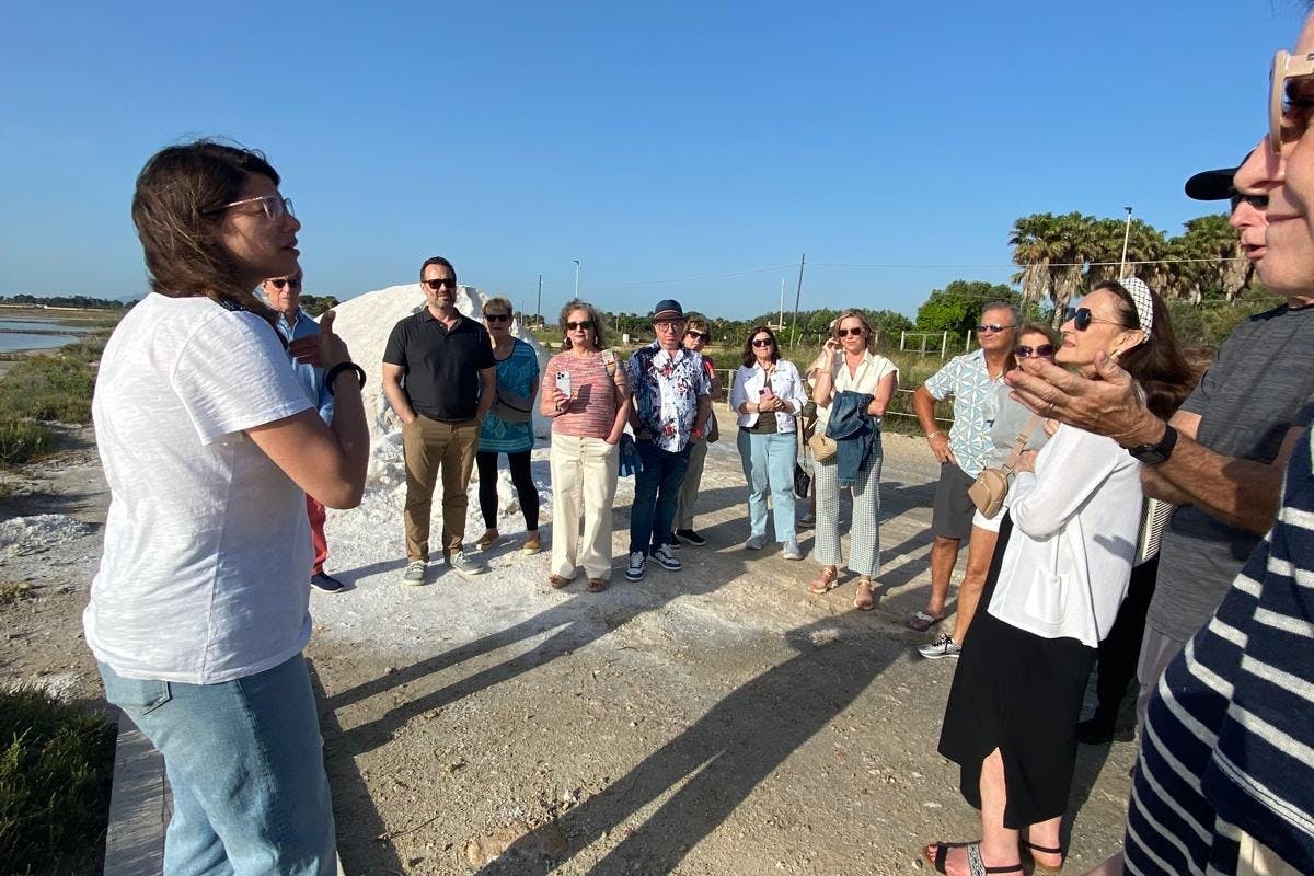 A group of people outdoors listening to a speaker on a sunny day, standing on a gravel path with greenery in the background.