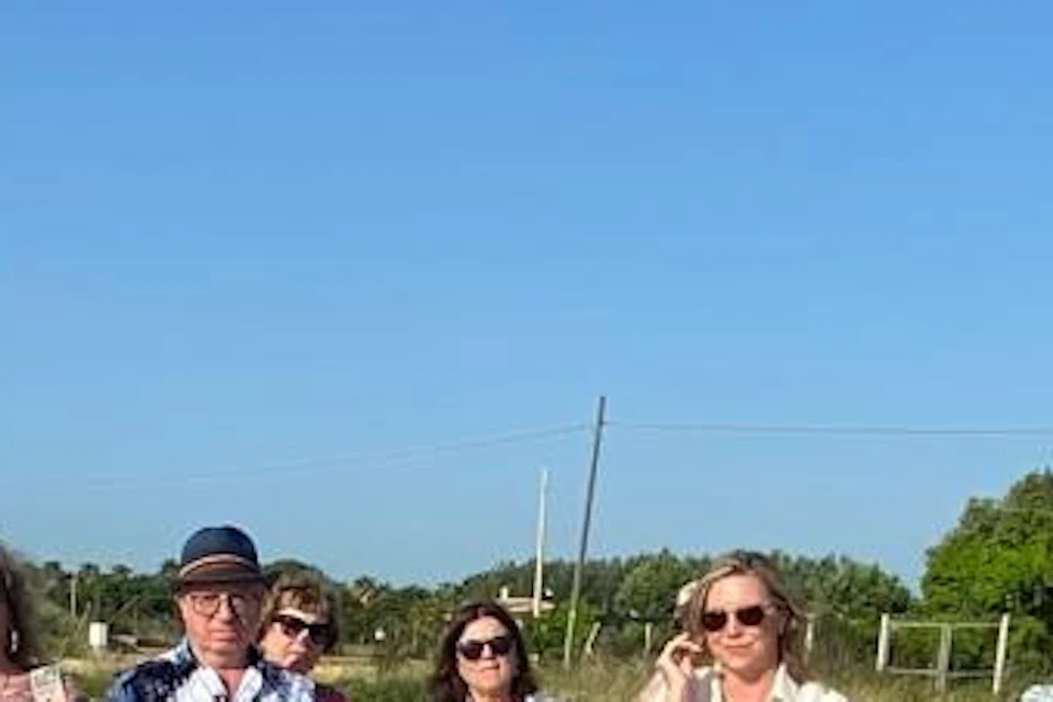 A group of people outdoors listening to a speaker on a sunny day, standing on a gravel path with greenery in the background.