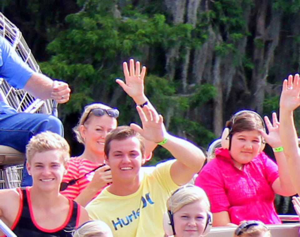 People on an airboat wave while gliding through wetlands, with lush trees and moss in the background.