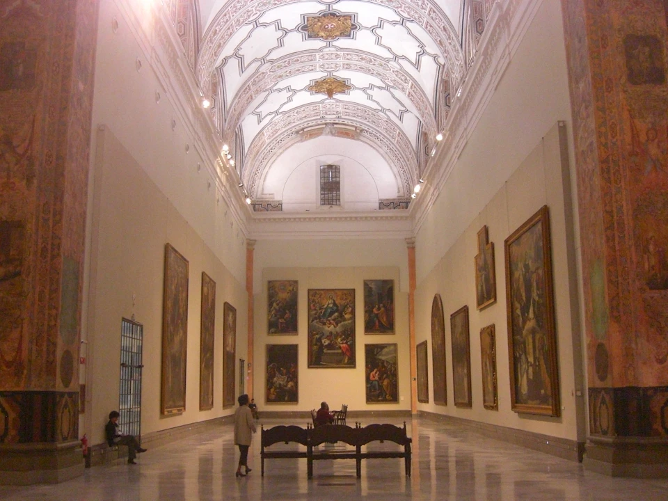 A grand hall in an art museum with ornate arches, large paintings on the walls, and a few people observing.