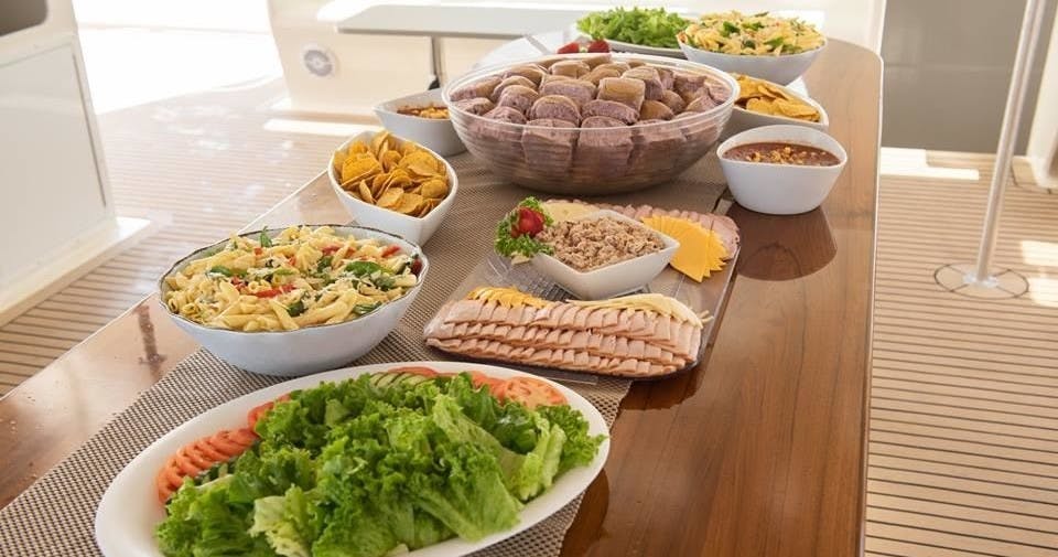 Uma mesa cheia de vários pratos, incluindo salada, carnes fatiadas, pãezinhos, massa, nachos e outros produtos alimentares.