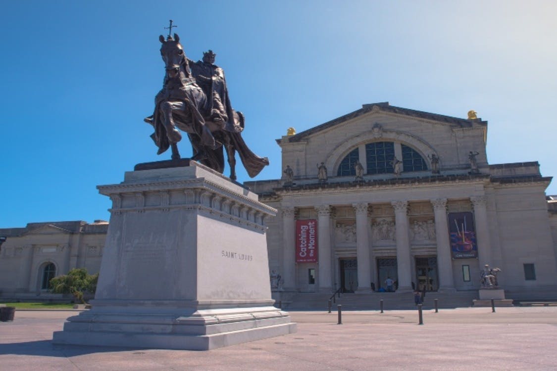 Ιππικό άγαλμα του Αγίου Λουδοβίκου μπροστά από ένα κλασικό κτίριο με μεγάλους κίονες και πανό. Καθαρός γαλάζιος ουρανός από πάνω.