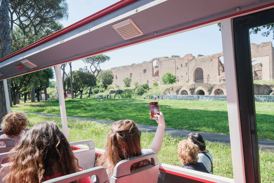 Tourists on a bus taking photos of ancient ruins in a grassy area under a clear sky.